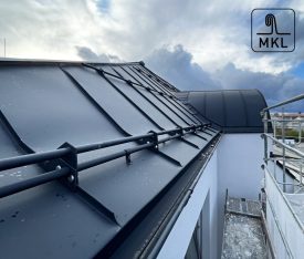 moderný bytový dom s antracitovou falcovanou strechou MKL, viacúrovňové strešné roviny, biela fasáda, sklenené balkóny, zamračené svetlo, realistická architektúra, presné detaily o